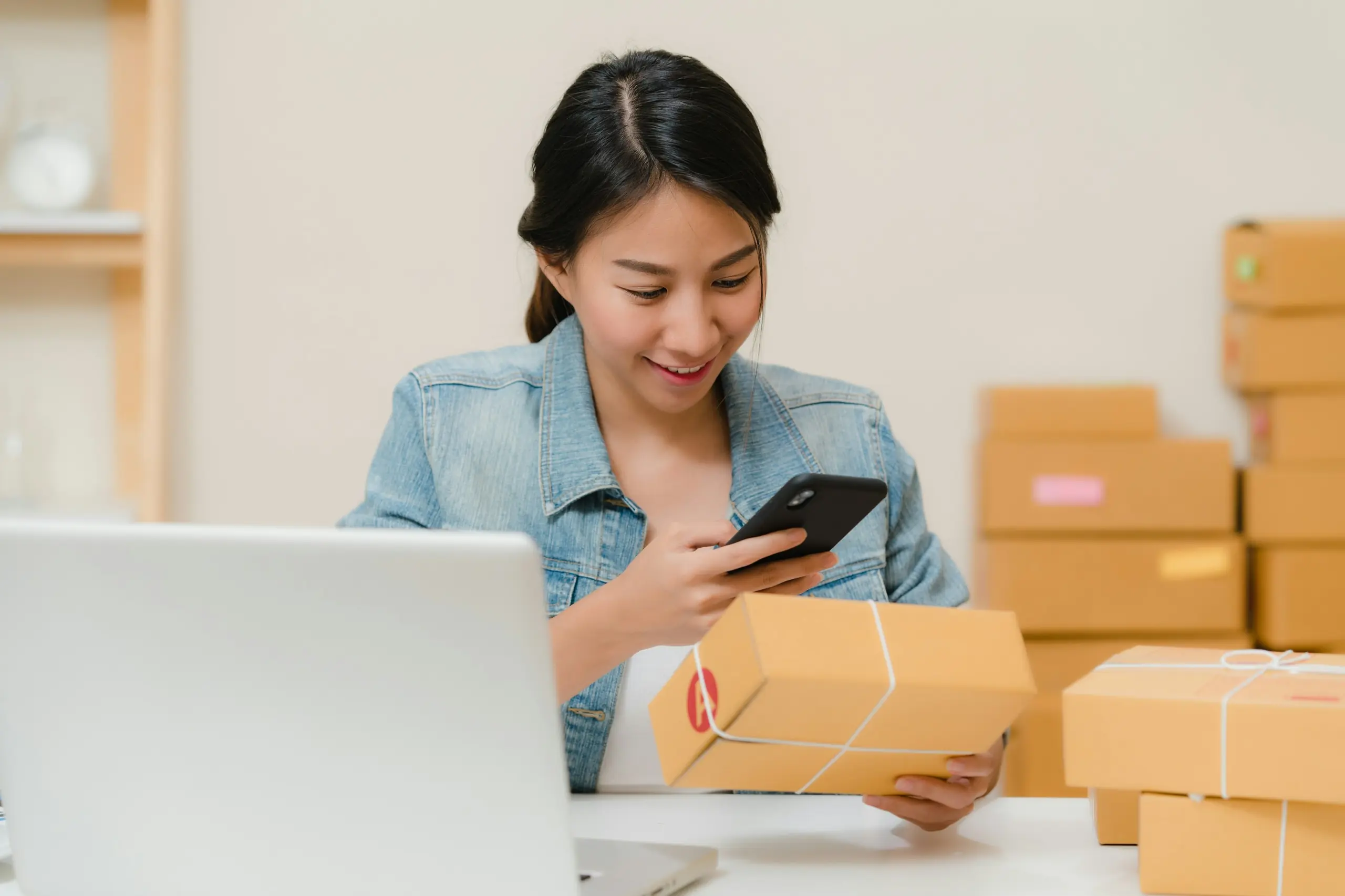 A seller at her desk checks a parcel against an order on her phone, with packed boxes ready to send.